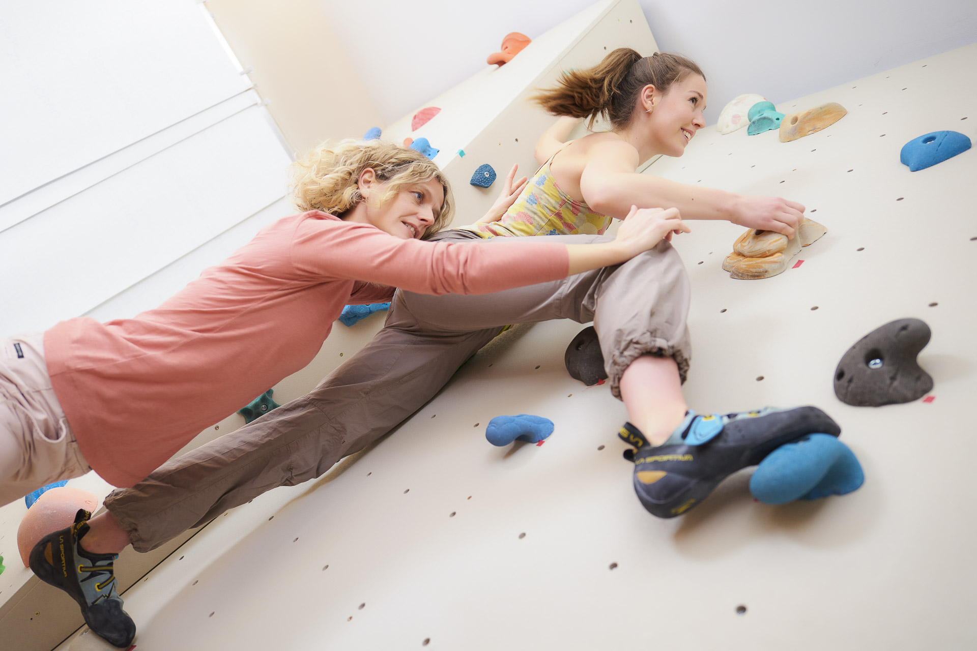 benedikt-kletterwand-3 eine junge fraun an einer bunten kletterwand unterstützt von einer frau in rosa pullover