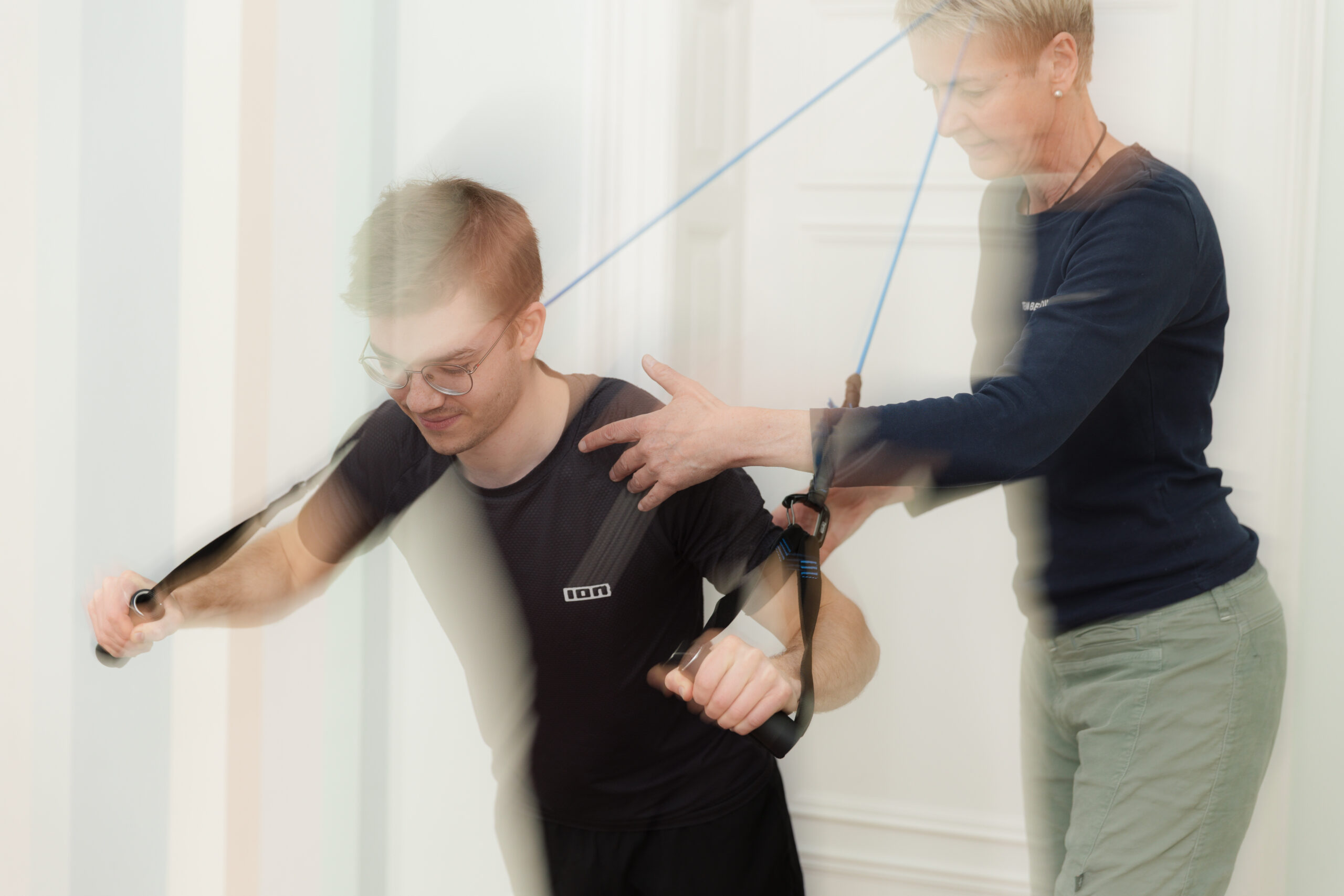 bettina-benedikt-physiotherapie-behandlung-theraband-praxis-benedikt-muenchen Bettina Benedikt führt Physiotherapie-Behandlung mit Theraband an Schulter und Arm eines jungen Patienten in der Praxis Benedikt München
