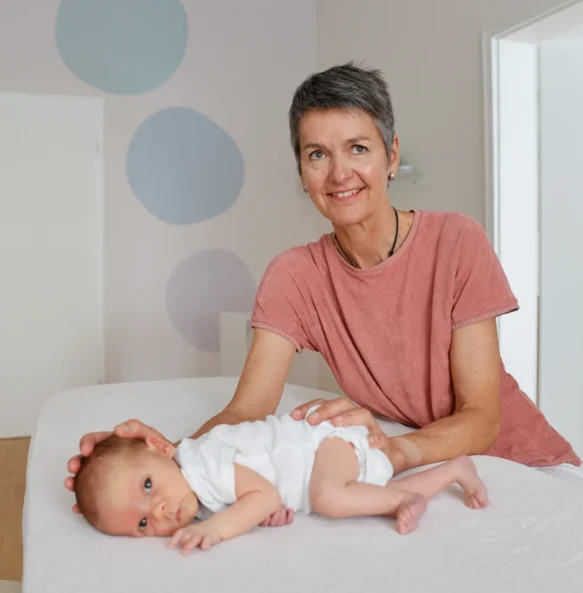 20250725-_F1A1101_compressed_2000px (1) Eine lächelnde Frau in rosa Shirt hält ein Baby auf weißem Bettlaken mit pastellblauen Kreisen im Hintergrund