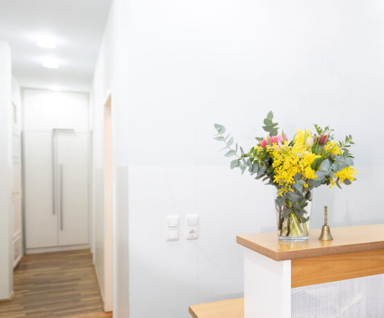 empfang-praxis-benedikt-muenchen-blumen-tresen Heller Empfangsbereich der Praxis Benedikt München mit Holztresen und buntem Blumenstrauß in Glasvase