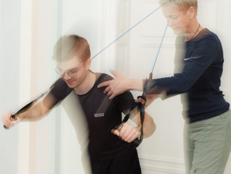 Bettina Benedikt führt Physiotherapie-Behandlung mit Theraband an Schulter und Arm eines jungen Patienten in der Praxis Benedikt München